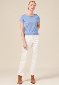 Top azzurro chiaro a maniche corte con vita aderente, abbinato a pantaloni bianchi a gamba dritta e stivaletti beige. Sfondo neutro.