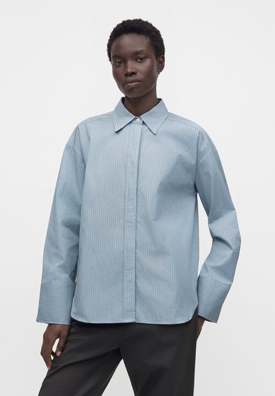 Personne à la peau foncée portant une chemise bleu clair à fines rayures boutonnée avec de larges poignets et un pantalon foncé, debout devant un fond blanc uni.