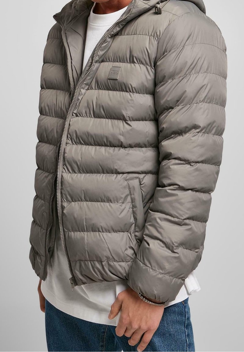 Man i en grå fodrad puffjacka med huva över vit skjorta och blå jeans, som står mot en neutral bakgrund.