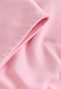 Lahka roza bombažna tkanina z mehkimi teksturami, prikazuje prepognjen rob s čistim šivom. Gladek površina, brez vidnih vzorcev ali okraskov.