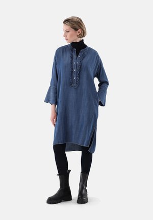 Denim-Tunika-Kleid mit Rüschenausschnitt und Knöpfen, langen Ärmeln, asymmetrischem Saum, getragen über schwarzen Leggings und schwarzen Stiefeletten.