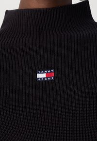 Gros plan sur un vêtement en maille côtelée noire avec un petit écusson rectangulaire Tommy Jeans en blanc, rouge et marine centré sur la poitrine.