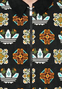Črna kordna jakna s središnjim srebrnim zadrgo, okrašena z repetitivnimi geometrijskimi cvetličnimi in kronastimi vzorci v oranžni, zeleni, beli in teal barvi.