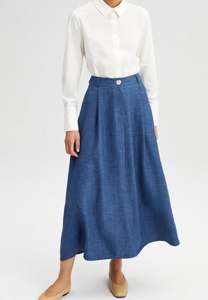 Femme portant un chemisier blanc à manches longues et une jupe midi bleue taille haute avec des chaussures plates beige, debout devant un fond uni.