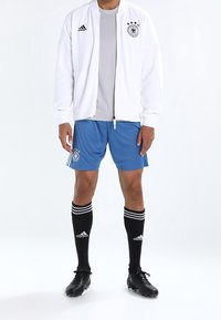 Bílá sportovní bunda přes šedé tričko, modré šortky, černé ponožky s bílými pruhy a černé fotbalové kopačky. Obsahuje německé fotbalové logo.