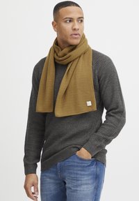 Brun strikket tørklæde draperet rundt om en grå tekstureret sweater, kombineret med blå jeans. Bemærkelsesværdig rektangulær tørklædeform og varierende strikkemønstre.