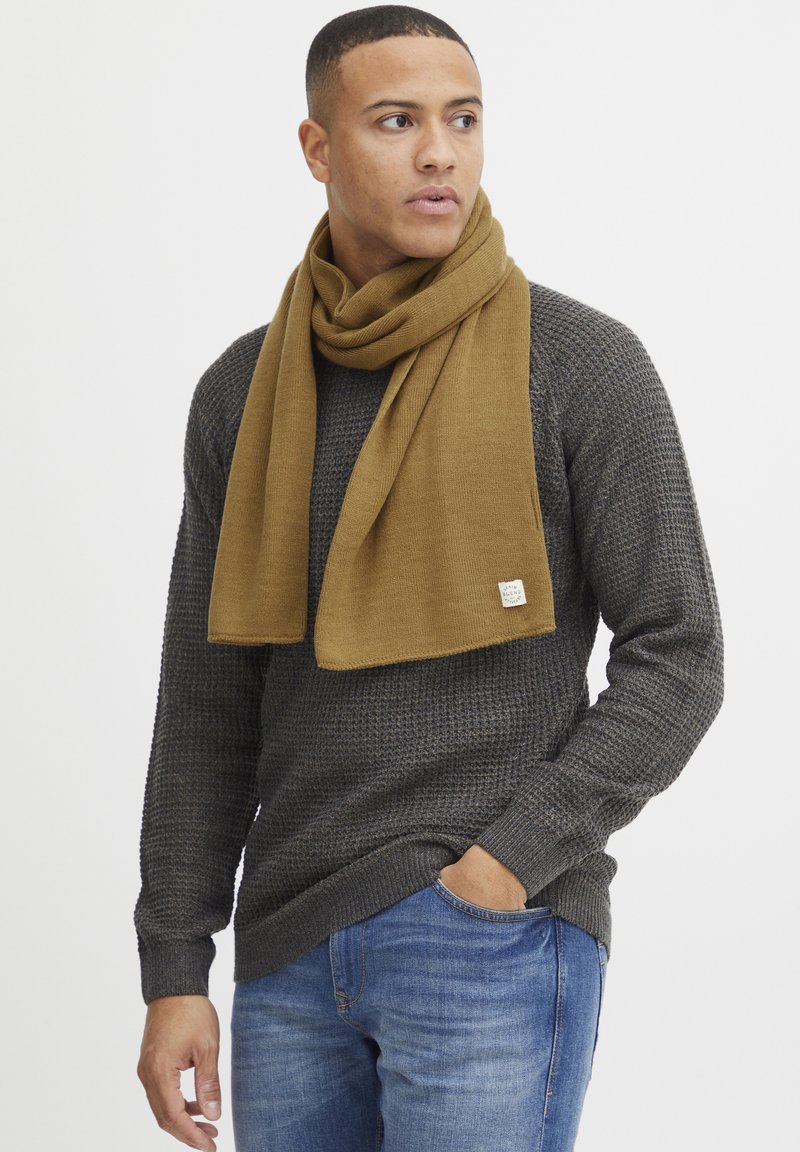 Brun strikket tørklæde draperet rundt om en grå tekstureret sweater, kombineret med blå jeans. Bemærkelsesværdig rektangulær tørklædeform og varierende strikkemønstre.