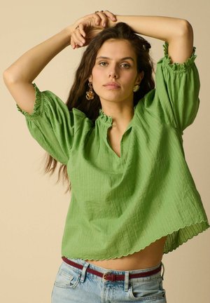 Femme aux longs cheveux bruns portant un chemisier vert texturé avec des bords festonnés et un jean bleu clair, posant avec les bras levés au-dessus de la tête.