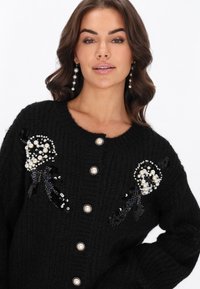 Czarny sweter z dzianiny z perłowymi i cekinowymi zdobieniami z przodu, okrągłym dekoltem i pięcioma zapięciami na guziki.