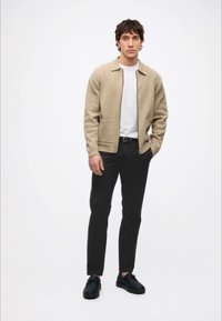 Mann steht und trägt eine beige Reißverschlussjacke, ein weißes T-Shirt, schwarze Hose und schwarze Schuhe vor einem einfarbigen weißen Hintergrund.