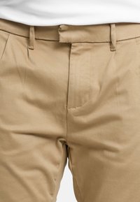 Primo piano di pantaloni beige con passanti per cintura indossati con una camicia bianca infilata, che evidenzia la vita anteriore e l'area di chiusura.