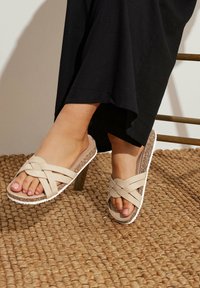 Des sandales à brides croisées beiges présentent une semelle intermédiaire en liège et une semelle extérieure blanche texturée, le tout mis en valeur sur un fond de tapis tissé naturel.