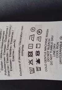 Textile care label featuring symbols for washing, drying, and ironing instructions. White fabric with printed black care icons and text.