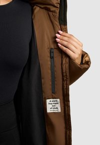 Giacca isolante marrone con foderatura nera, dotata di una tasca con zip e di un'etichetta che recita "Un abbraccio caldo fatto di stoffa."