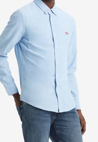 Camisa de botón azul claro hecha de algodón, con collar y mangas largas. Presenta un pequeño parche de logo rojo en el pecho. Combinada con jeans de mezclilla.