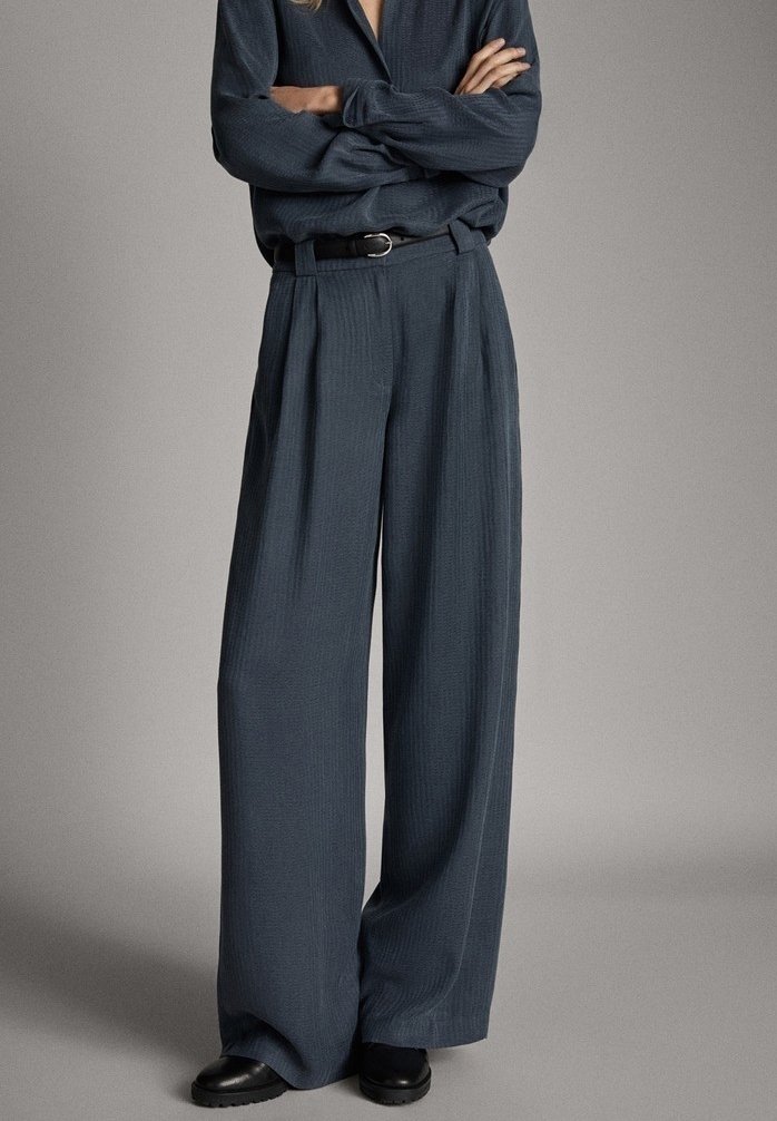 Femme portant un pantalon large gris foncé, un haut à manches longues assorti avec col, ceinture noire et chaussures noires, debout avec les bras croisés.