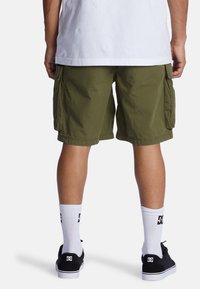 Pantalones cortos de carga verdes con bolsillos laterales, usados con calcetines blancos y zapatillas negras. La tela parece texturizada y ligera.