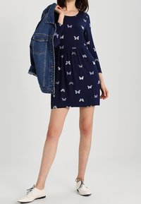 Mörkblå klänning med långa ärmar, prydd med fjärils mönster i olika pastellfärger, bärs med en jeansjacka och vita skor.