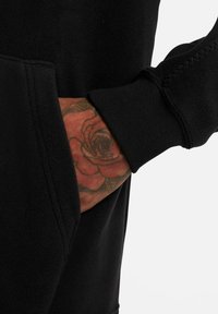 Schwarzer Fleece-Pullover mit Kängurutasche und gerippten Bündchen. Ein Tattoo einer Rose ist auf der Hand sichtbar, die in der Tasche liegt.