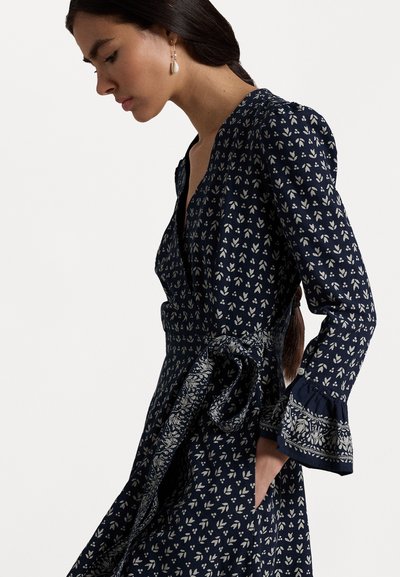 Tummansininen mekko, jossa on valkoinen kukkakuvio, V-pääntie, pitkät hihat ja levenevät hihansuut. Läpiveto vyötärö ja solmittava vyöyksityiskohta.