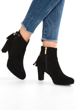 Bottes chevilles en daim noir avec talons carrés et fermetures éclair latérales, portées avec un jean bleu à ourlet effiloché.