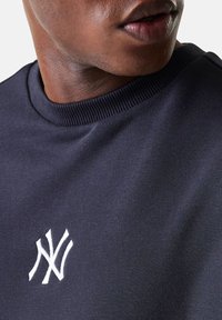 Primer plano de una persona que lleva una sudadera azul oscuro con un logo de NY bordado en blanco cerca del cuello.