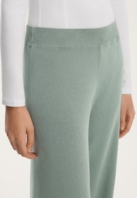 Žebrované světle zelené kalhoty s elastickým pasem, zobrazené spolu s bílým dlouhým rukávem. Hladká textura, přiléhavý střih.