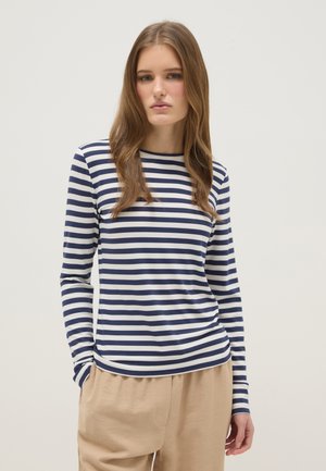 Jonge vrouw met lang bruin haar, gekleed in een marineblauw-wit gestreept shirt met lange mouwen en beige broek, staand met handen in de zakken.