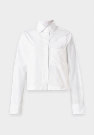 Donna che indossa una camicia bianca abbottonata infilata in pantaloni beige a gamba larga, con le mani nelle tasche, in piedi davanti a uno sfondo semplice.