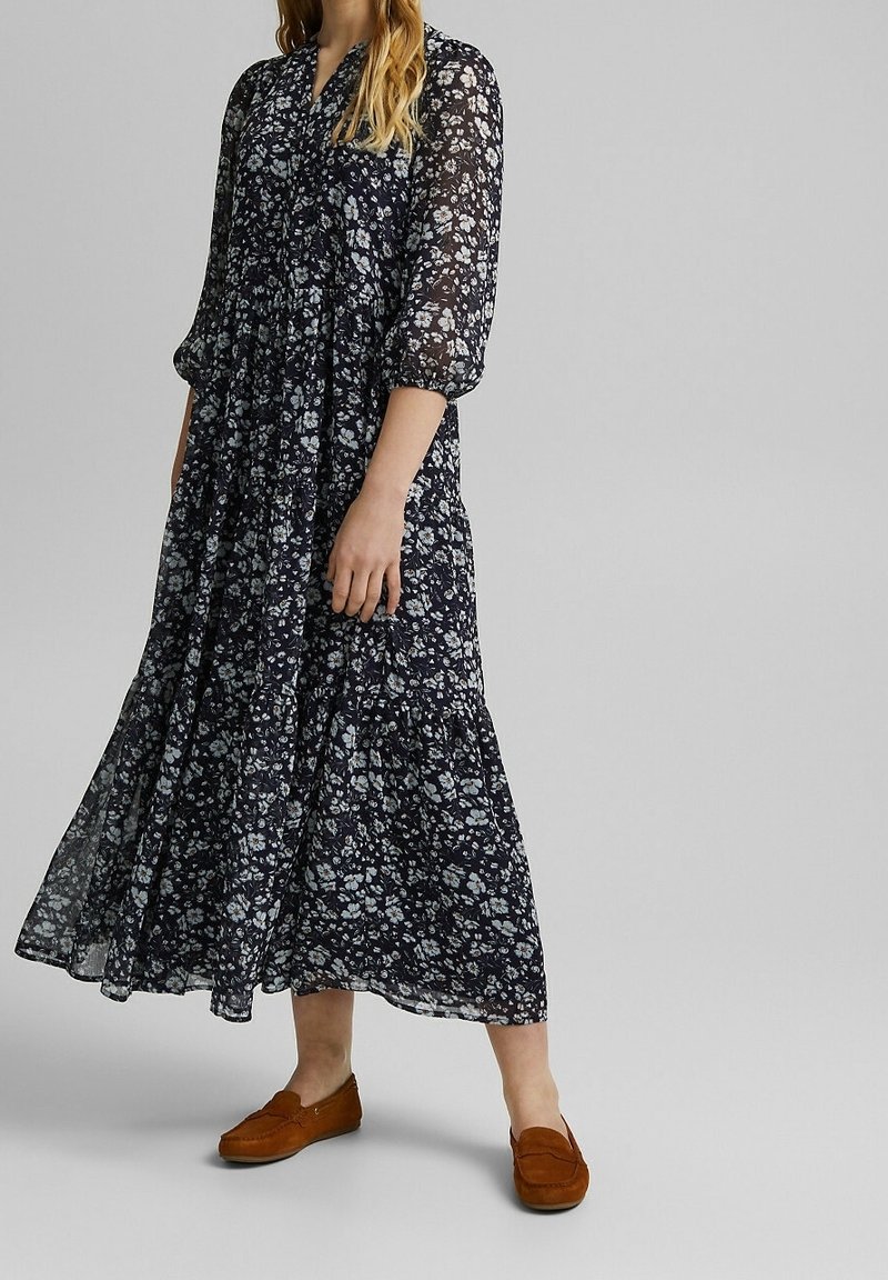 Femme portant une robe midi noire à fleurs avec des manches ballon et des mocassins marron, debout devant un fond gris uni.