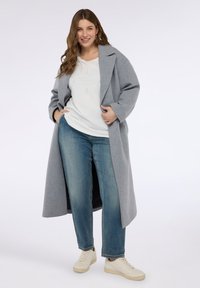 Kobieta uśmiechająca się, ubrana w długi szary płaszcz na białą koszulę, niebieskie dżinsy i białe trampki, stojąca na tle jasnego, jednolitego tła.