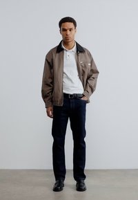 Καφέ ελαφρύ μπουφάν με μαύρο γιακά πάνω από λευκό πόλο πουκάμισο, συνδυασμένο με σκούρα μπλε τζιν και μαύρα παπούτσια, στέκεται μπροστά σε απλό φόντο.