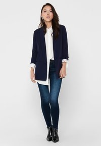 Blazer bleu marine à coupe cintrée, à un bouton sur le devant et deux poches, superposé sur une chemise blanche et associé à un jean en denim foncé et des bottes noires.