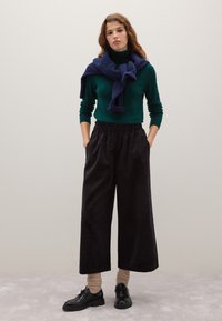 Maglione dolcevita verde scuro, sciarpa blu navy, pantaloni neri a gamba larga e scarpe nere. Presenta un tessuto testurizzato e una vita elasticizzata.
