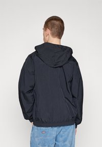 Young man viewed from behind wearing a black hooded windbreaker jacket and light blue denim jeans with back pockets.