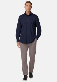 Muž stojí s rukami v kapsách, oblečený v námornícky modrej košeli s dlhým rukávom na gombíky, svetlohnedých nohaviciach a čiernych teniskách s bielymi podrážkami.