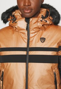 Φουσκωτό μπουφάν σε μεταλλικό μπρονζέ με μαύρες ρίγες, διαθέτοντας κουκούλα με επένδυση από γούνα, τσέπες με φερμουάρ και κεντημένο λογότυπο στο στήθος.