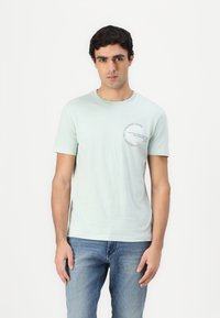 T-shirt verde chiaro a girocollo con una piccola grafica circolare sull'area sinistra del petto, realizzata in un tessuto morbido con una texture liscia.