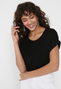 Top nero a maniche corte con scollatura rotonda, tessuto liscio e maniche arrotolate, indossato con jeans bianchi e orecchini d'argento pendenti.
