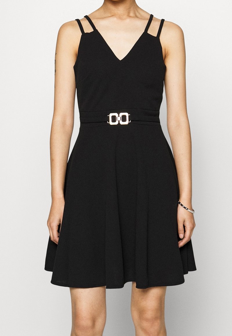 Femme portant une robe noire sans manches avec doubles bretelles et ceinture à boucle dorée, debout devant un fond uni.