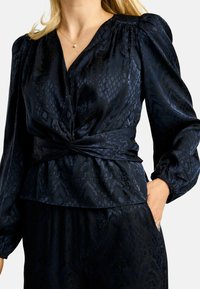 Tumesinine pluus, millel on tekstuuriga madu muster, pehme satiinist kangas, V-kaelus, kogutud volangvarrukad ja vööümber sõlmitud detail.