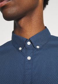 Camicia blu navy con bottoni e motivo strutturato, con colletto aperto e bottoni bianchi. Vista ravvicinata del colletto e dello scollo.