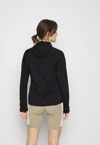 Sort letvægtsjakke med hætte, der har en glat overflade og tætsiddende design. Kombineret med beige shorts med sidelommer.