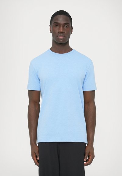 T-shirt en coton bleu à manches courtes, col rond et finition légèrement texturée. Design simple, coupe droite, porté avec un pantalon noir.