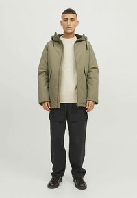 Blago zelen puhovka z kapuco, z zadrgo spredaj in stranskimi žepi. Oblečena preko krem puloverja in črnih cargo hlač. Črni čevlji.