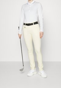 Biała longsleeve polo z kołnierzykiem i guziczkami, żółte spodnie, czarny pasek z logo, białe trampki i kij golfowy. Gładka faktura.