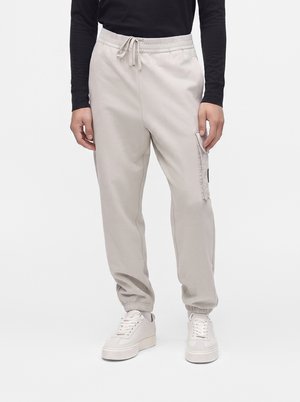 La personne porte un pantalon de jogging cargo beige clair avec des poignets élastiques et une taille à cordon, associé à des baskets blanches et un haut noir à manches longues.