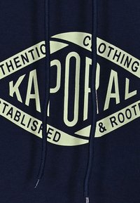 Sudadera azul marino de algodón, con un gran estampado gráfico en verde claro que incluye el texto "KAPORAL" y detalles adicionales de la marca. Con dos cordones.