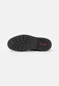 Suola di scarpa in gomma nera con scanalature profonde e robuste per una migliore aderenza. Presenta una texture in rilievo e un piccolo dettaglio con logo rosso.