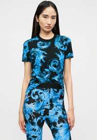 Νεαρή γυναίκα ντυμένη με μαύρη φόρμα που ταιριάζει, με φωτεινά μπλε στροβιλισμένα υδάτινα σχέδια, στέκεται μπροστά σε απλό φωτεινό φόντο.